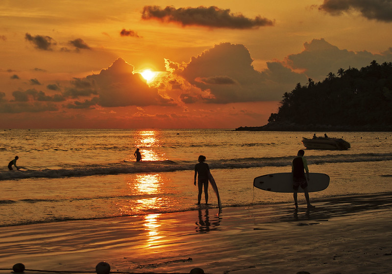 Zachód słońca na Kata Beach na Phuket, z surferami i złotym światłem odbijającym się w wodzie.