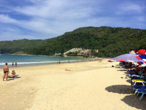 Plaża Nai Harn na Phuket – szeroka, piaszczysta plaża z turkusową wodą, otoczona zielonymi wzgórzami, popularna wśród turystów i surferów.