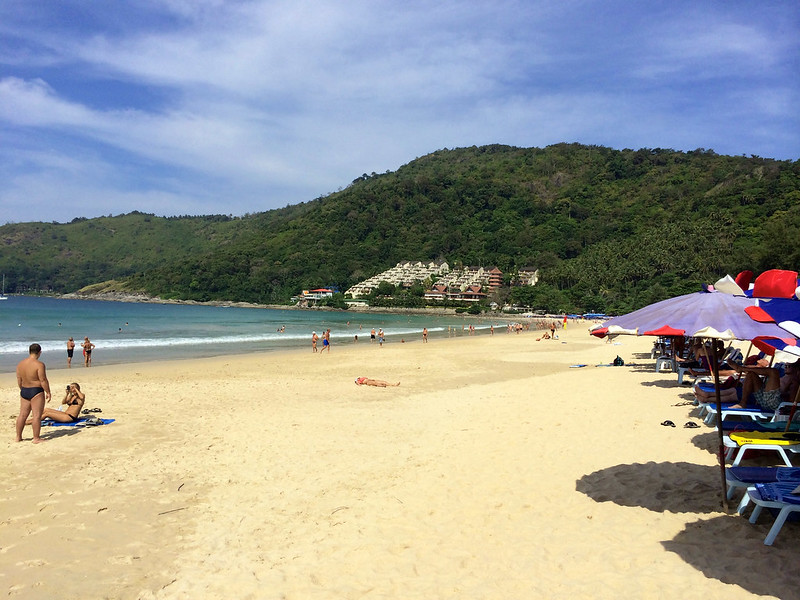 Plaża Nai Harn na Phuket – szeroka, piaszczysta plaża z turkusową wodą, otoczona zielonymi wzgórzami, popularna wśród turystów i surferów.