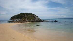 Spokojna plaża Ya Nui na Phuket – widok na piaszczyste wybrzeże z turkusową wodą i skałą w tle, o zachodzie słońca.