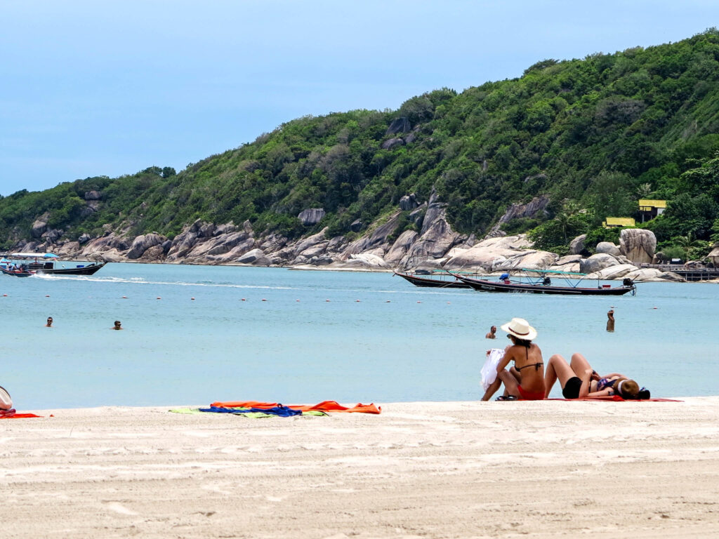 Widok na plażę Haad Rin na Koh Phangan z białym piaskiem, turkusową wodą i zielonymi wzgórzami w tle.