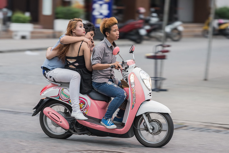 bieta i mężczyzna jadą na skuterze w Tajlandii, ubrani w letnie ubrania, z uśmiechem na twarzy.